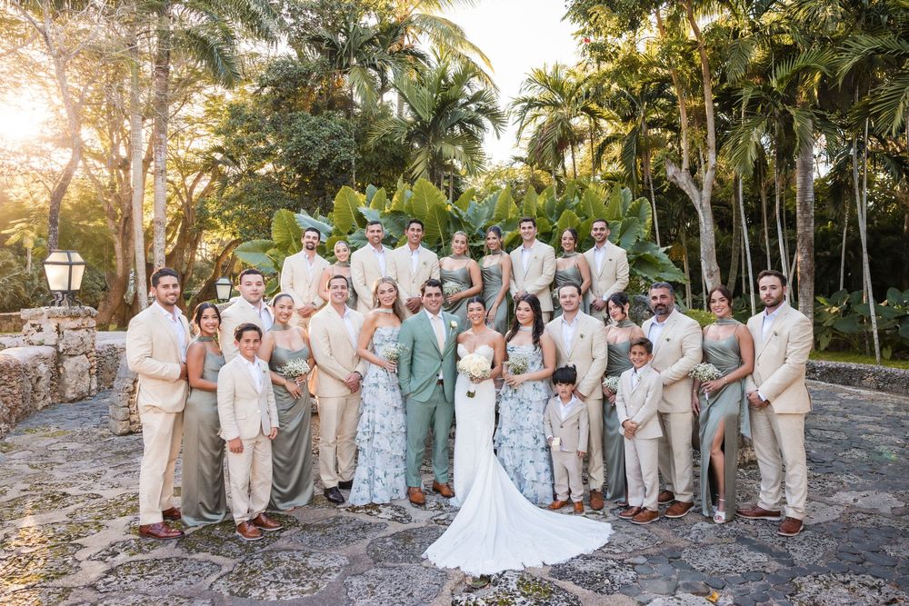 Wedding party poses in neutral and sage green attire against a tropical garden backdrop during sunset.