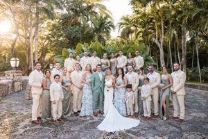 Wedding party poses in neutral and sage green attire against a tropical garden backdrop during sunset.