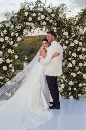 Romantic wedding couple embraces under a beautiful white floral arch at sunset in an outdoor garden setting.