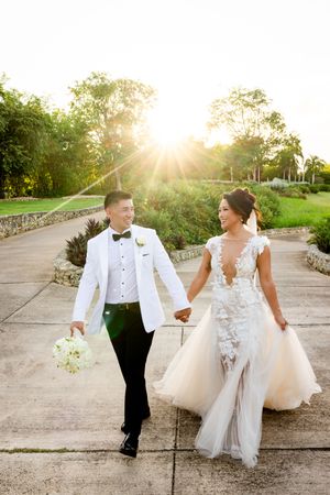 Bride and groom walk hand-in-hand at sunset, she in lace gown, he in white tuxedo jacket, on a garden pathway.