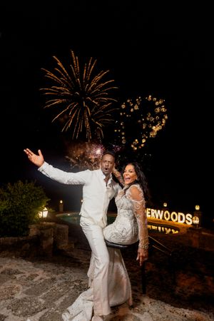 Couple in white wedding attire celebrating with fireworks and heart-shaped lights at an outdoor nighttime reception.