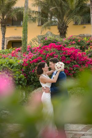 A couple embraces and dances in a garden setting with vibrant pink bougainvillea flowers and palm trees in the background. Sanctuary