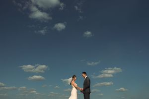 Wedding couple stands on a beach at sunset while a photographer captures their special moment.