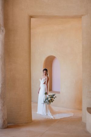 A person in an elegant white wedding gown holds a floral bouquet while standing in an archway with beige stone walls.
