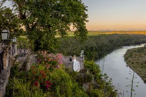 Scenic overlook at sunset shows a winding river below with stone railings and flowering plants in the foreground.
