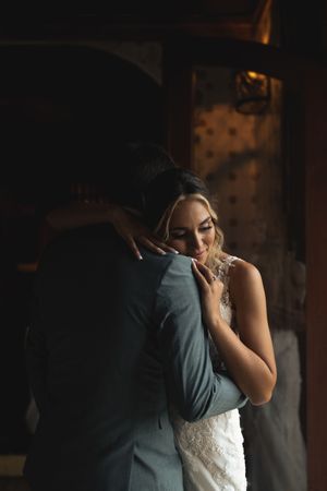 A tender moment between a couple embracing in a dimly lit space, with the bride wearing an elegant white lace gown.