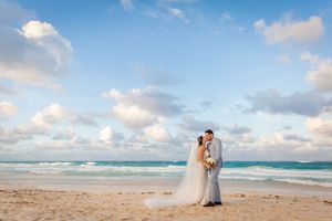 Couple embraces on sandy beach with turquoise ocean waves and white clouds in background during sunset.