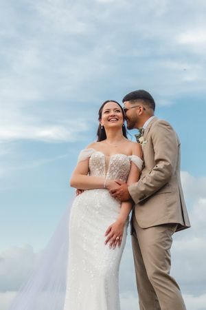 A couple shares a romantic moment outdoors, with the bride in a white lace gown and groom in a tan suit against blue sky.