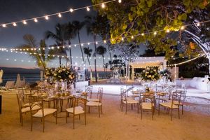 String lights illuminate an outdoor beach wedding reception setup with wooden chairs and tables under palm trees at dusk.
