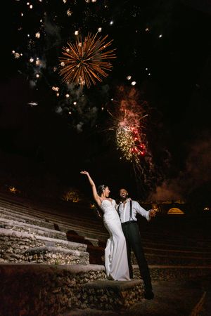 A couple sits on stone steps at night while colorful fireworks burst in the dark sky above them.