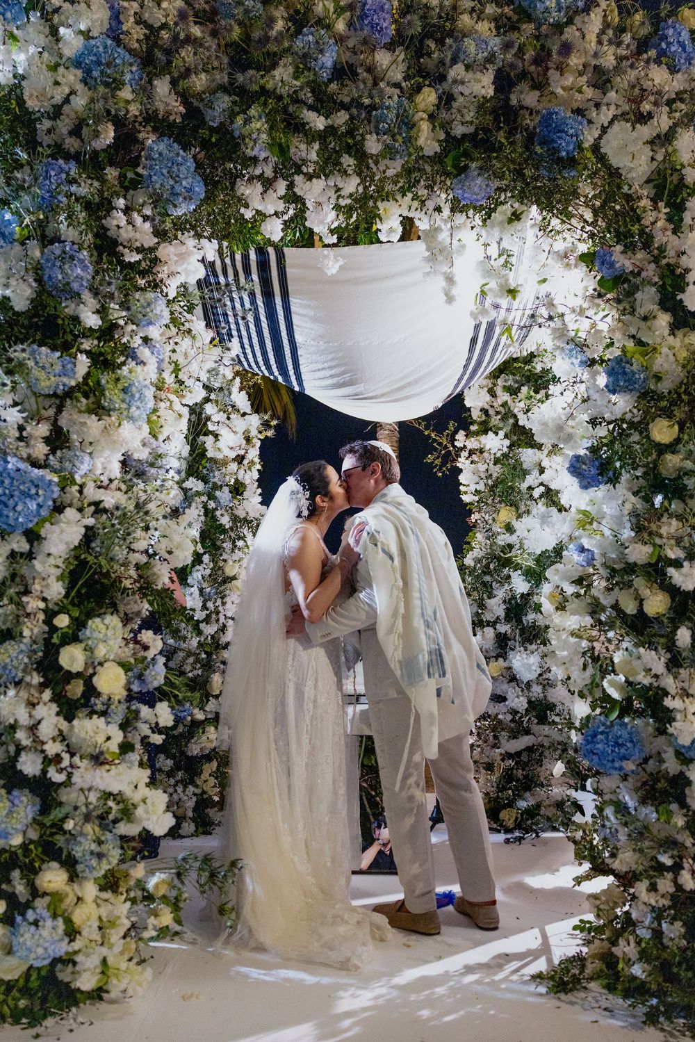 Bride and groom kissing under a floral chuppah adorned with blue and white hydrangeas at a Jewish wedding ceremony.