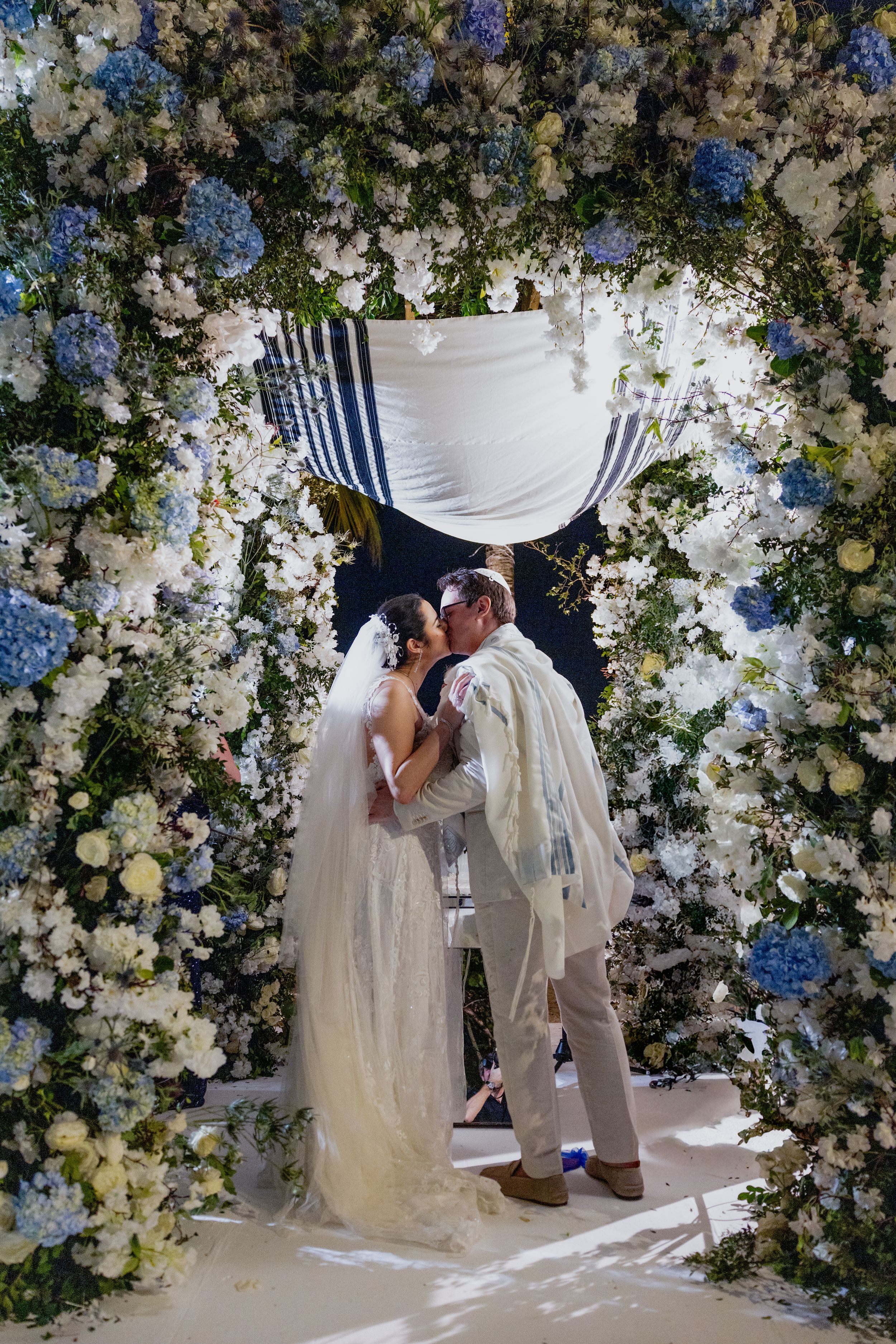 Bride and groom kissing under a floral chuppah adorned with blue and white hydrangeas at a Jewish wedding ceremony.