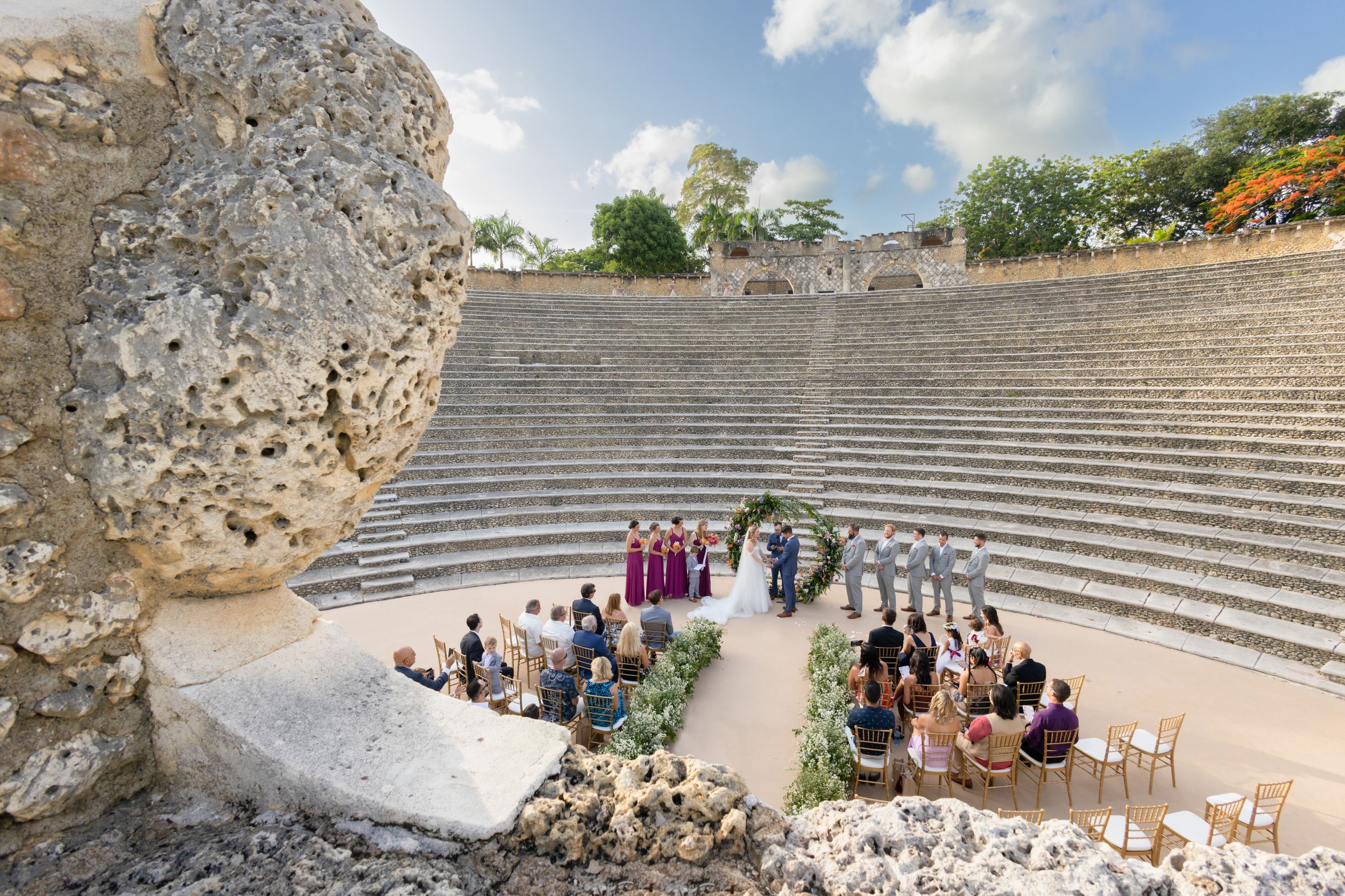 Ancient stone amphitheater steps and ruins under blue sky with scattered clouds and visitors exploring the historic site.