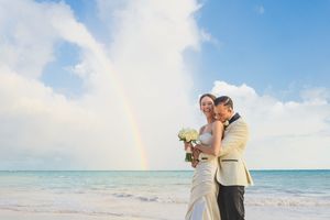 A couple embraces on a tropical beach with a rainbow in the background during a wedding ceremony. Sanctuary Cap Cana