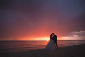Silhouetted figures stand together against a vibrant orange and purple sunset sky over the ocean horizon.