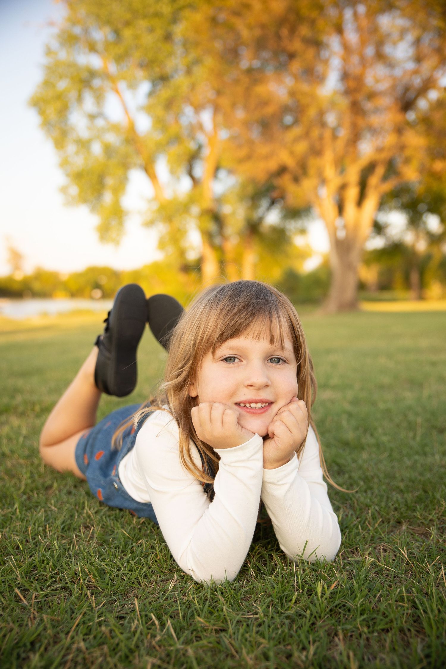 A child lies on autumn grass in a white sweater during golden hour with a playful expression.