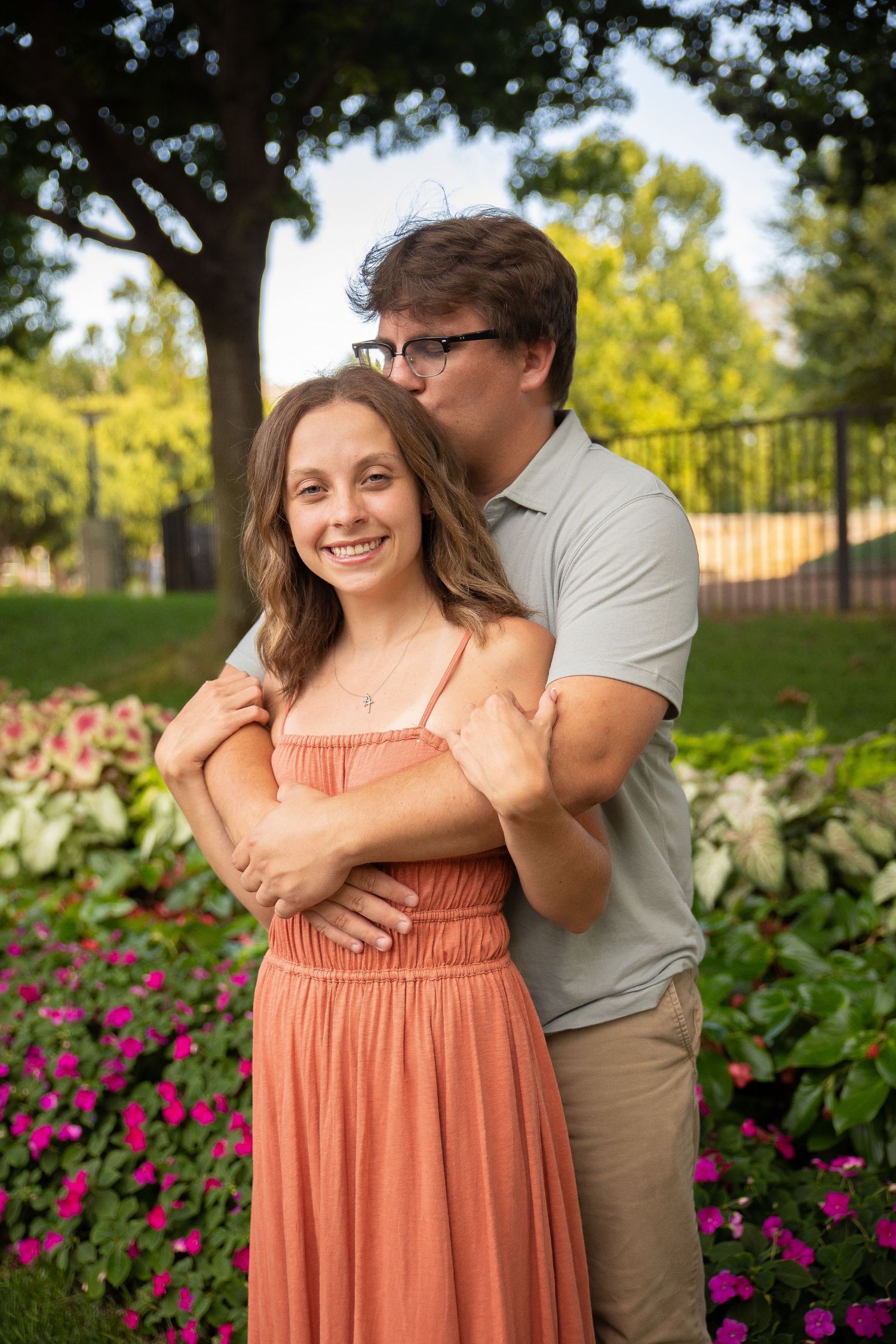 A couple embraces in a garden with vibrant purple flowers and greenery on a sunny afternoon.