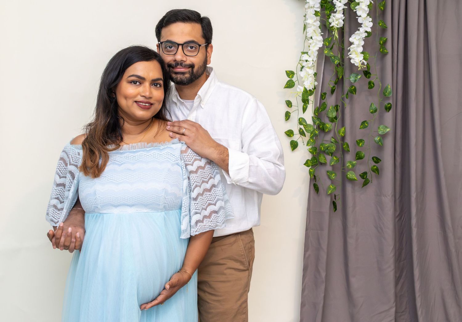 Expecting couple in blue and white outfits posing against floral backdrop indoors.