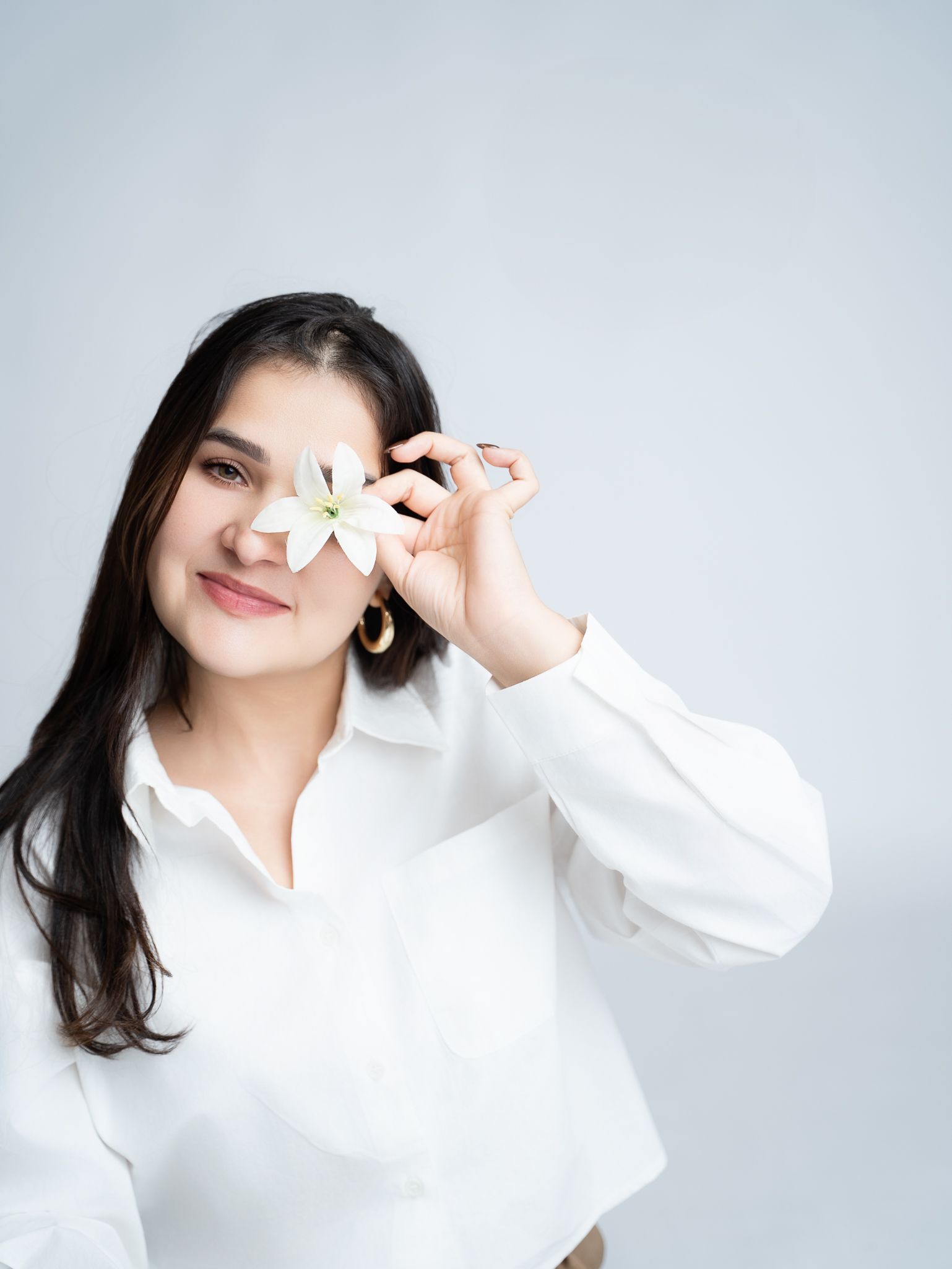 Person in white long sleeve top holds a white flower in front of their face against light gray background.