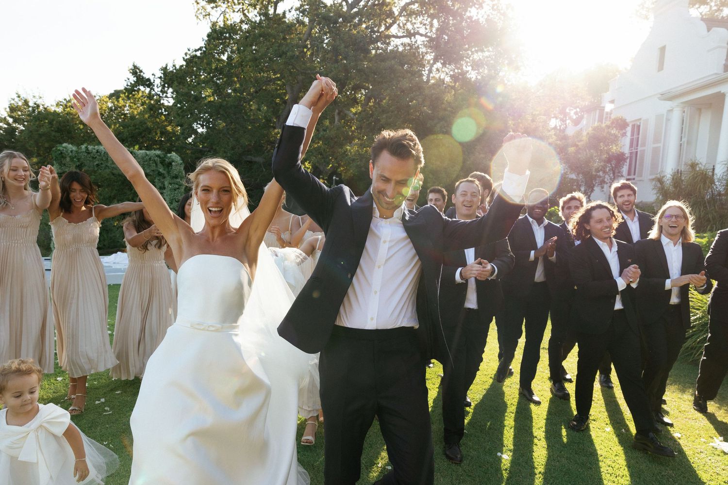 A wedding party jumps joyfully in unison on a sunny lawn during golden hour celebrations.