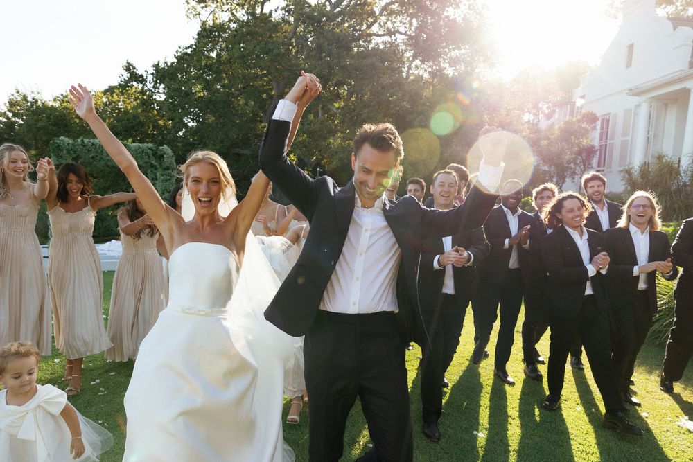 A wedding party jumps joyfully in unison on a sunny lawn during golden hour celebrations.