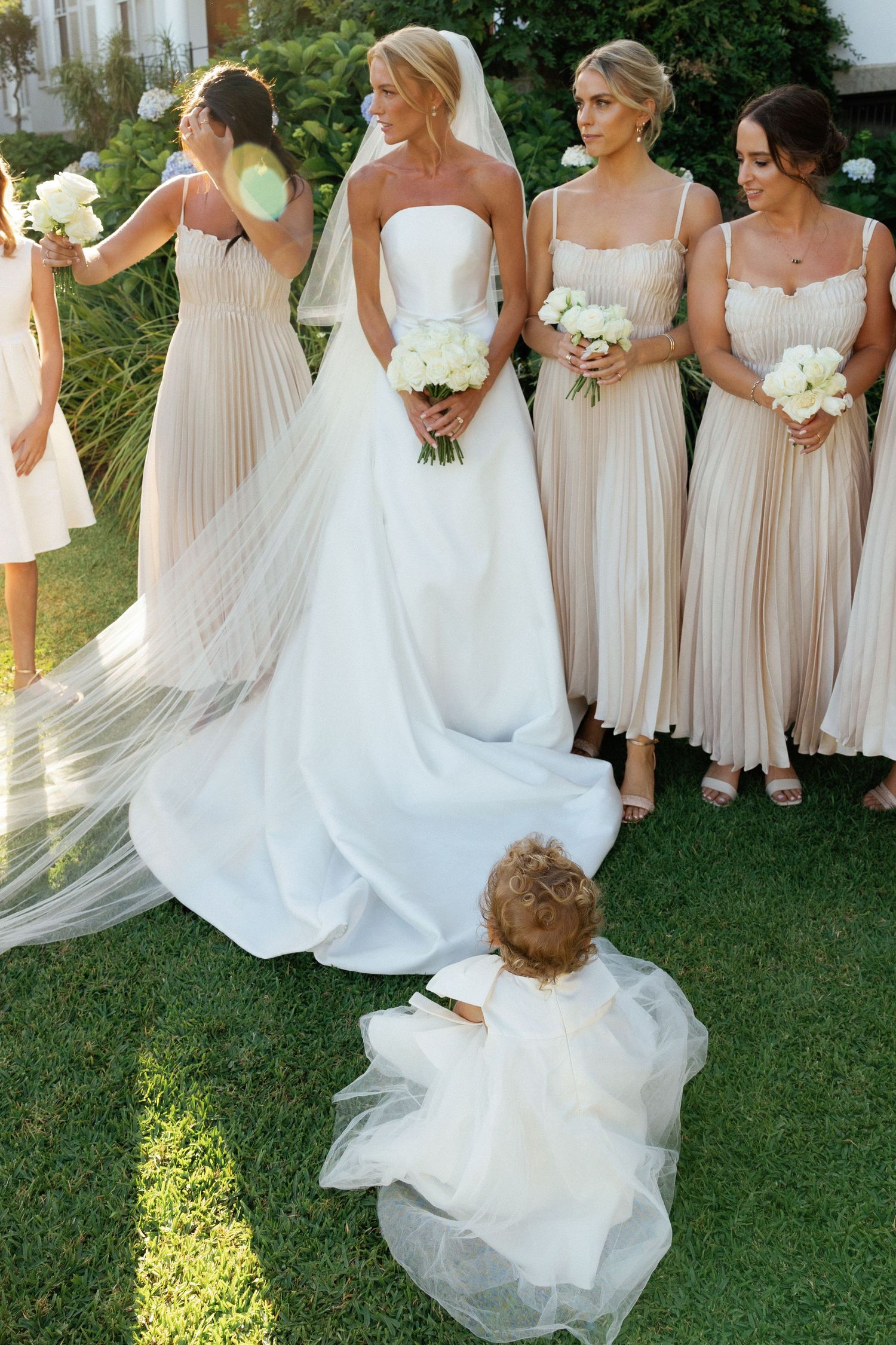 A bridal party in blush pink and white dresses poses together on a sunny lawn during an outdoor wedding celebration.