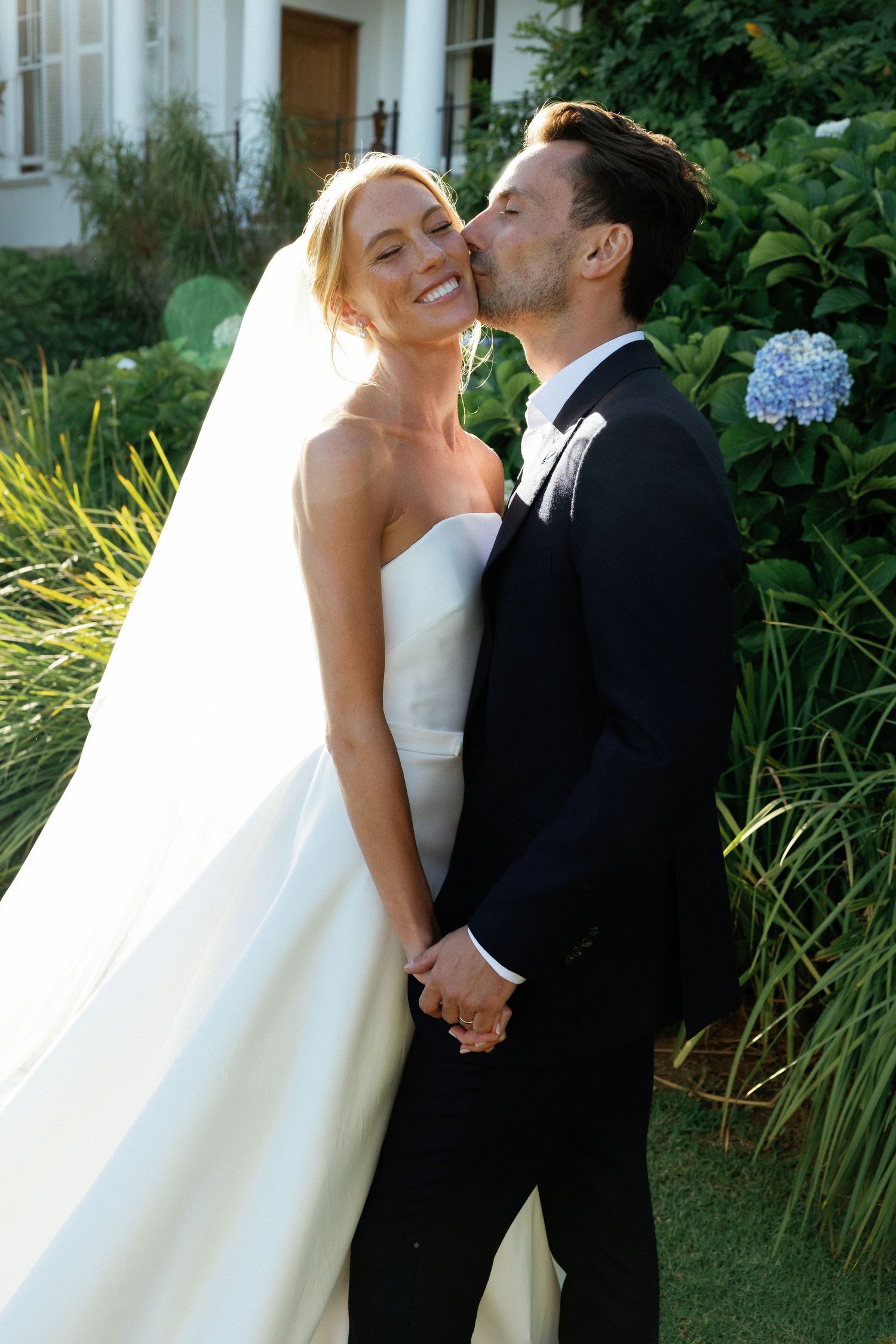 Newlyweds share a joyful moment together outdoors with lush greenery and yellow flowers in the background.