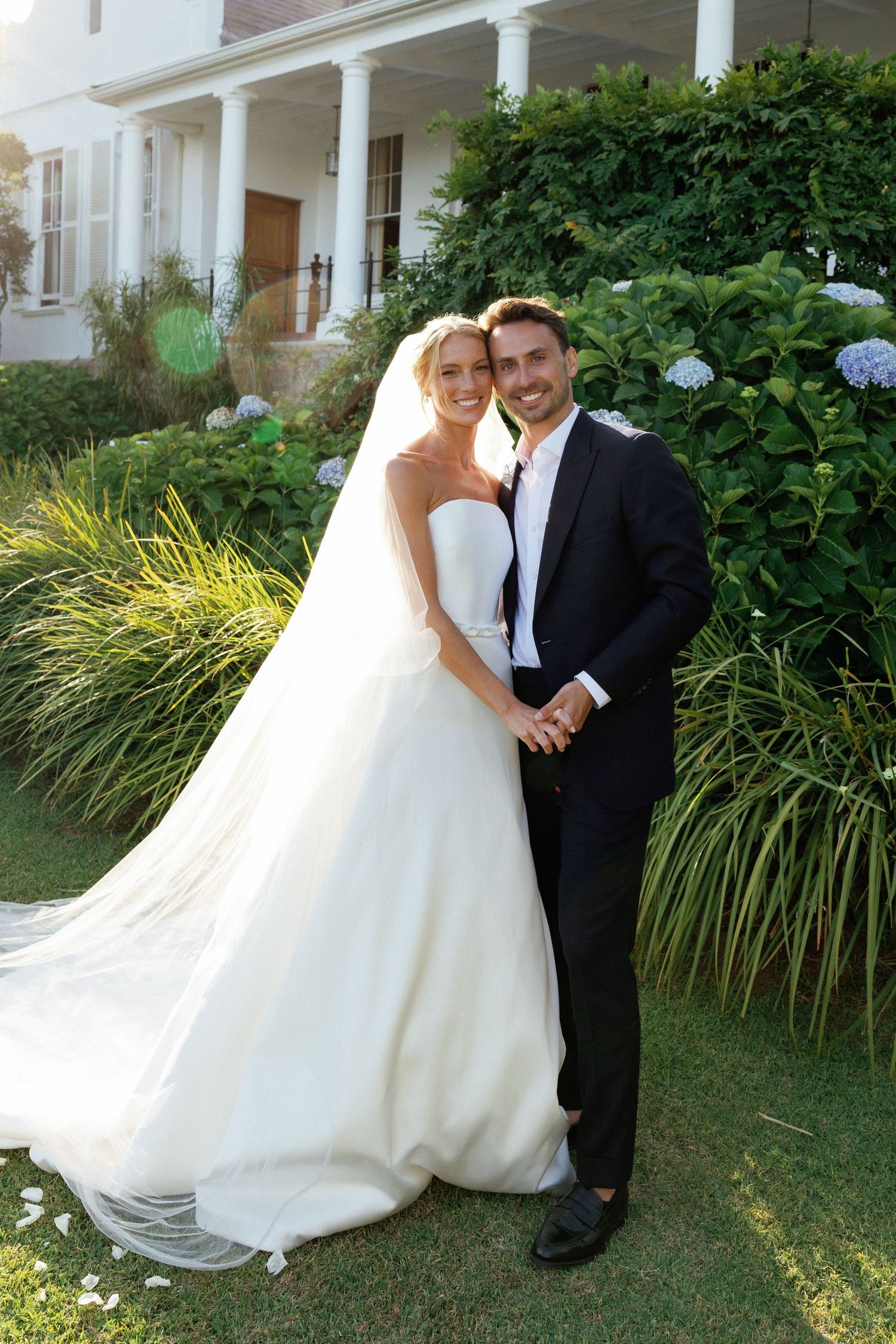 A bride in a flowing white wedding gown and groom in black suit pose together in a garden with hydrangeas and ornamental grasses.