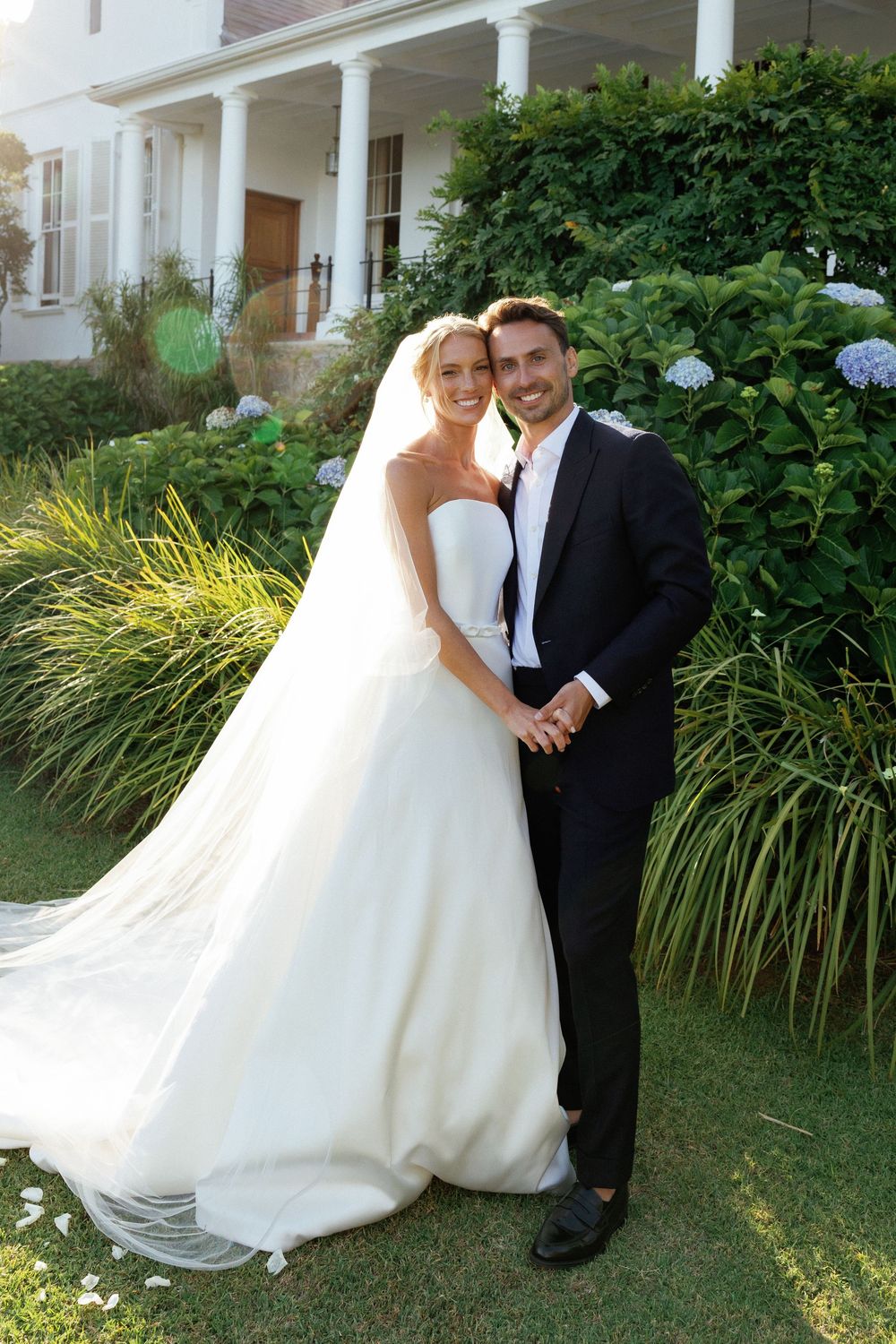 A bride in a flowing white wedding gown and groom in black suit pose together in a garden with hydrangeas and ornamental grasses.