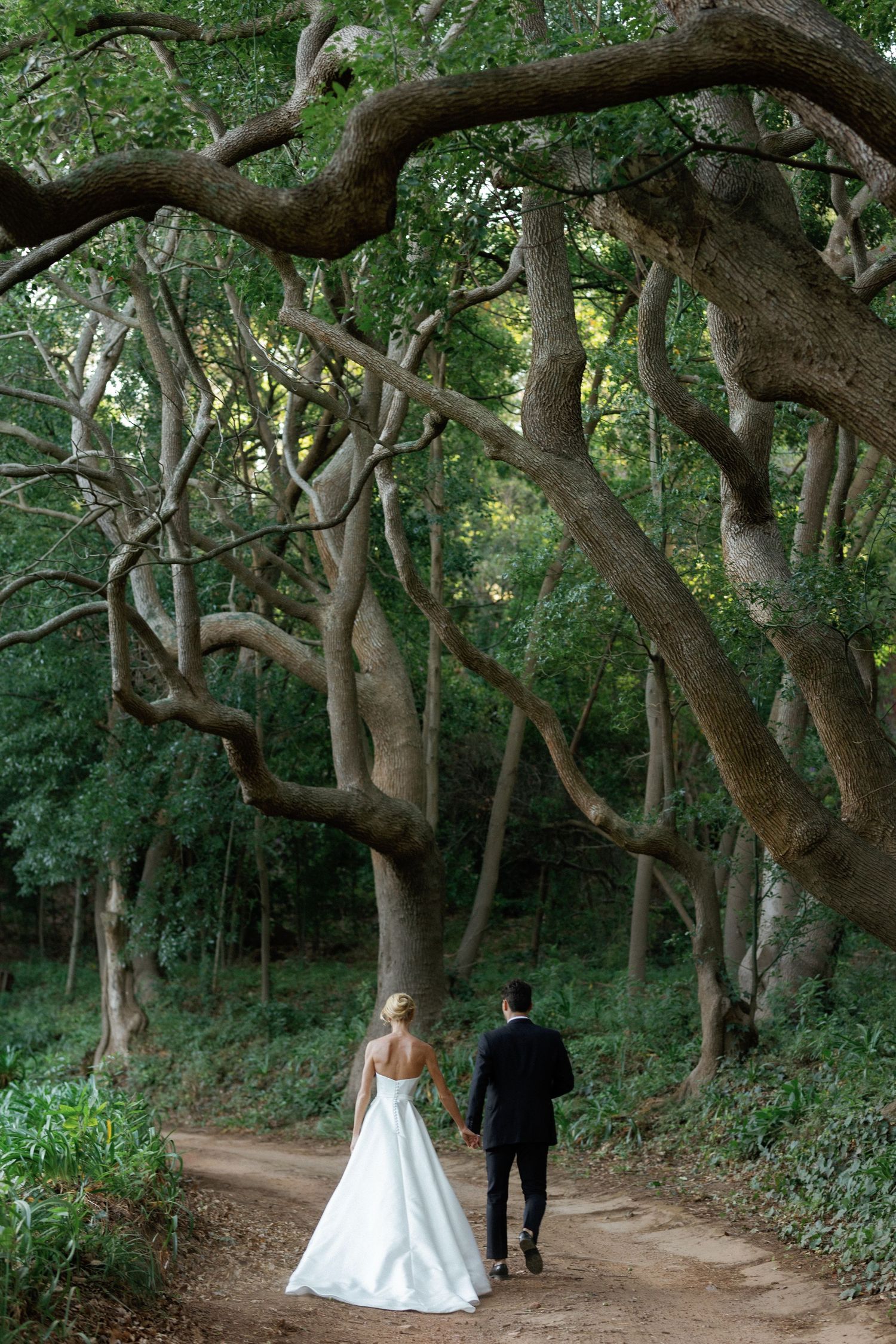A couple in wedding attire walks along a winding tree-lined path through a lush green forest setting.