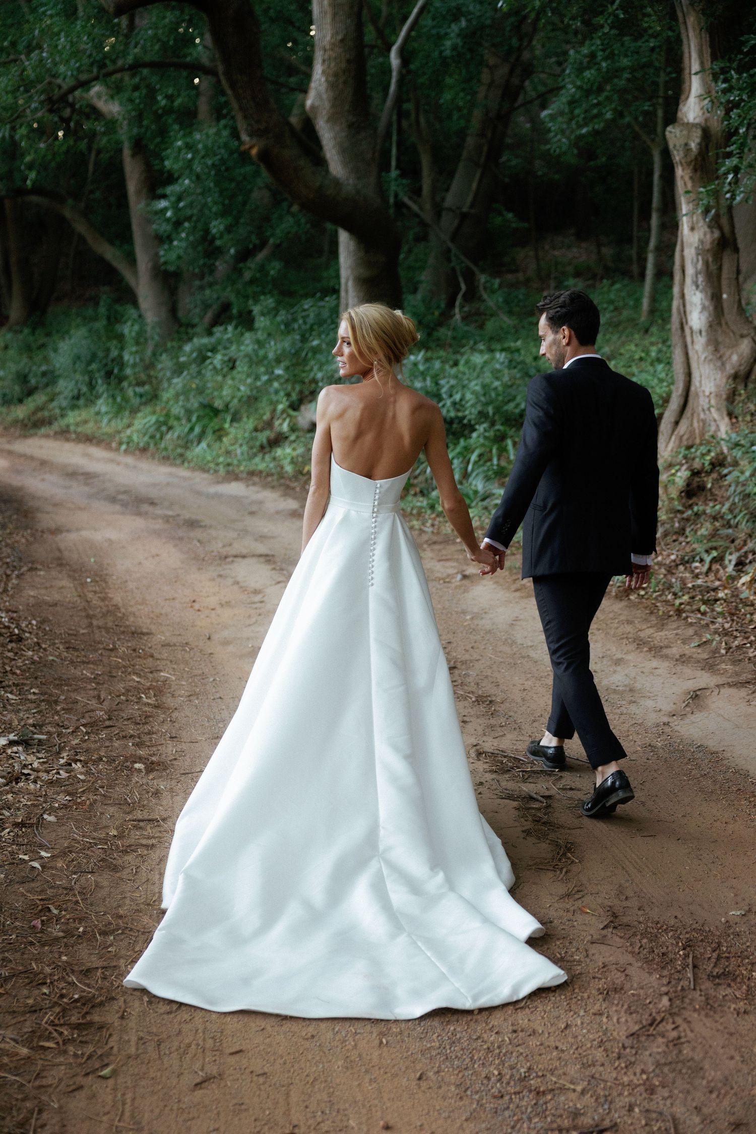A bride and groom walk along a winding dirt path through a wooded area during their wedding photo session.