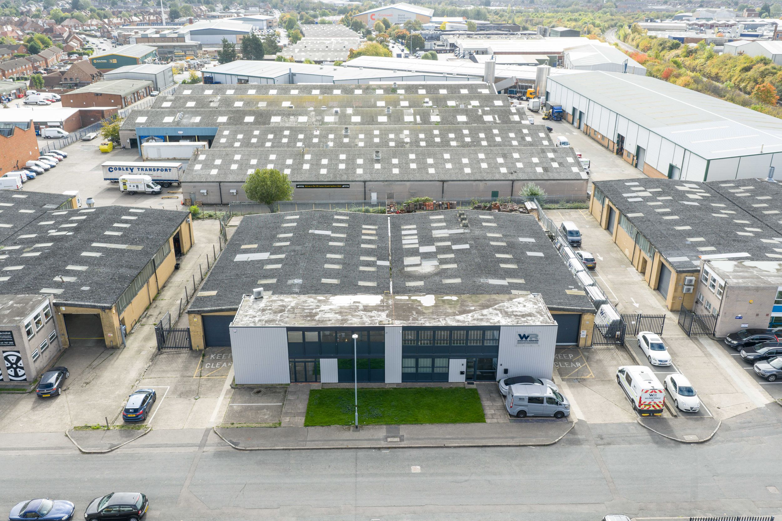 Aerial view of industrial warehouse buildings and parking lots with vehicles scattered throughout the complex.
