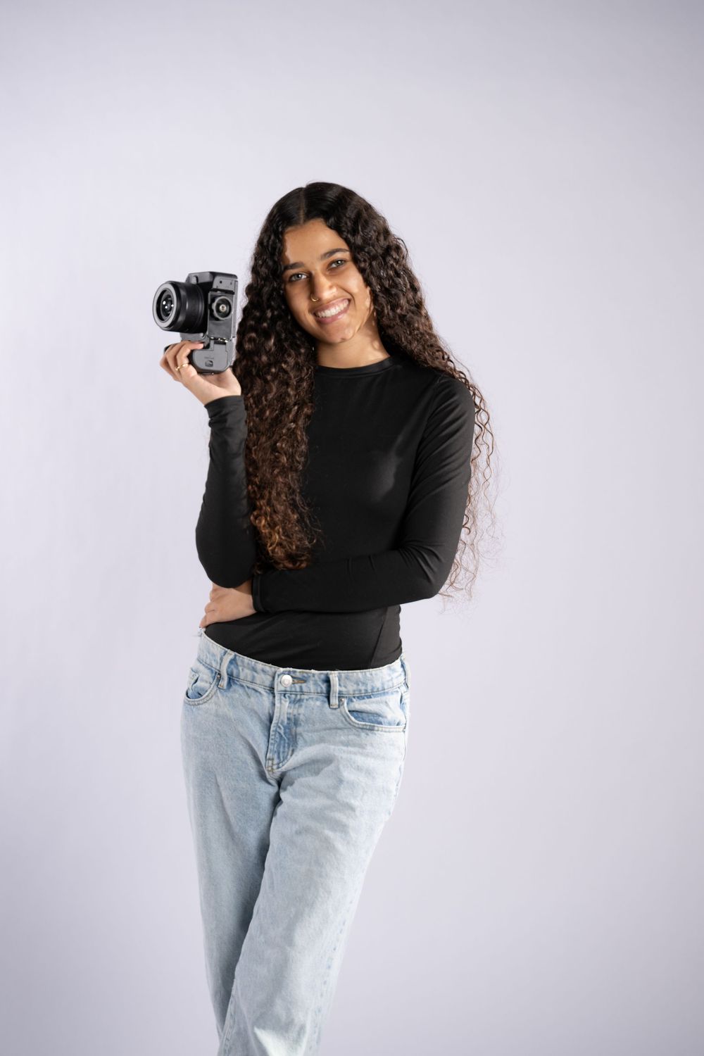 A photographer in light jeans and black top poses with a vintage camera against white background.
