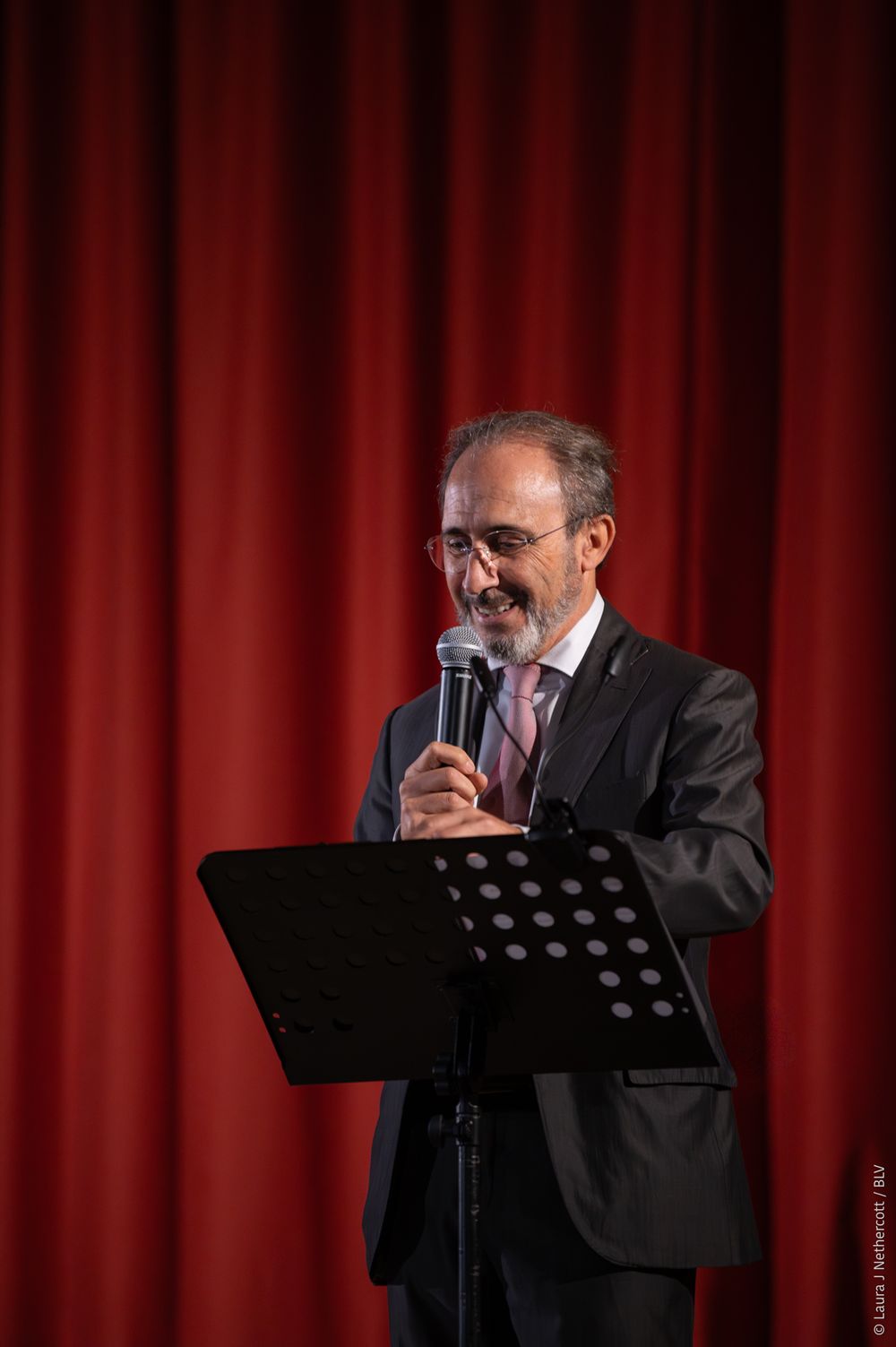 A speaker in a dark suit and glasses stands at a podium with a microphone against a red curtain backdrop.