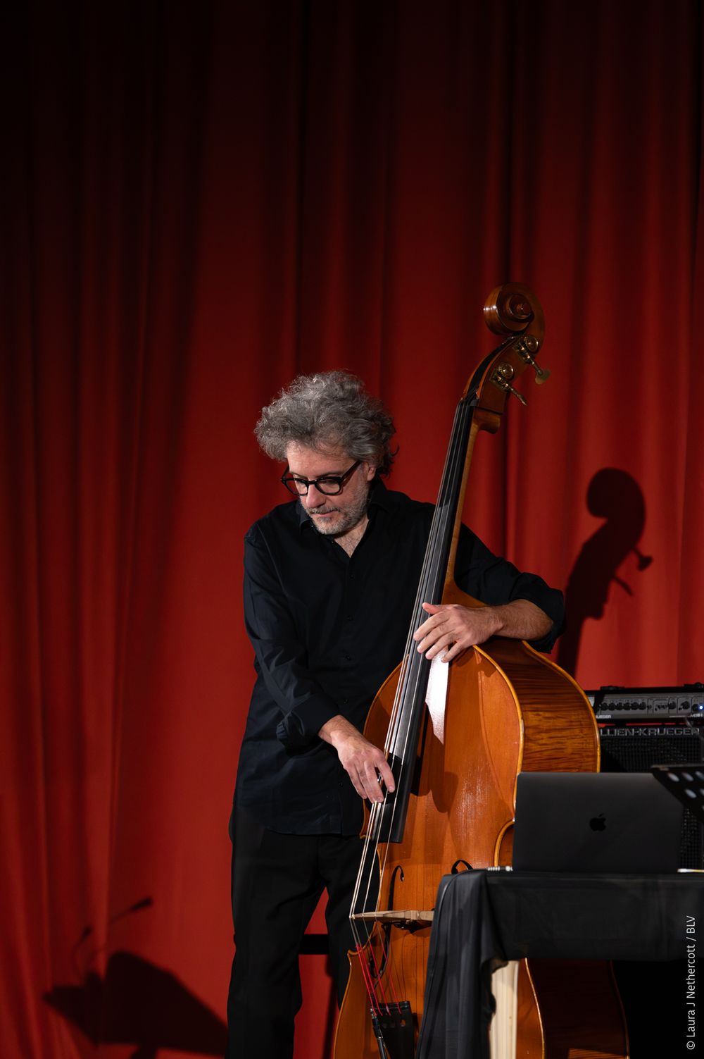 Musician in black attire performing with a double bass against a red curtain backdrop.