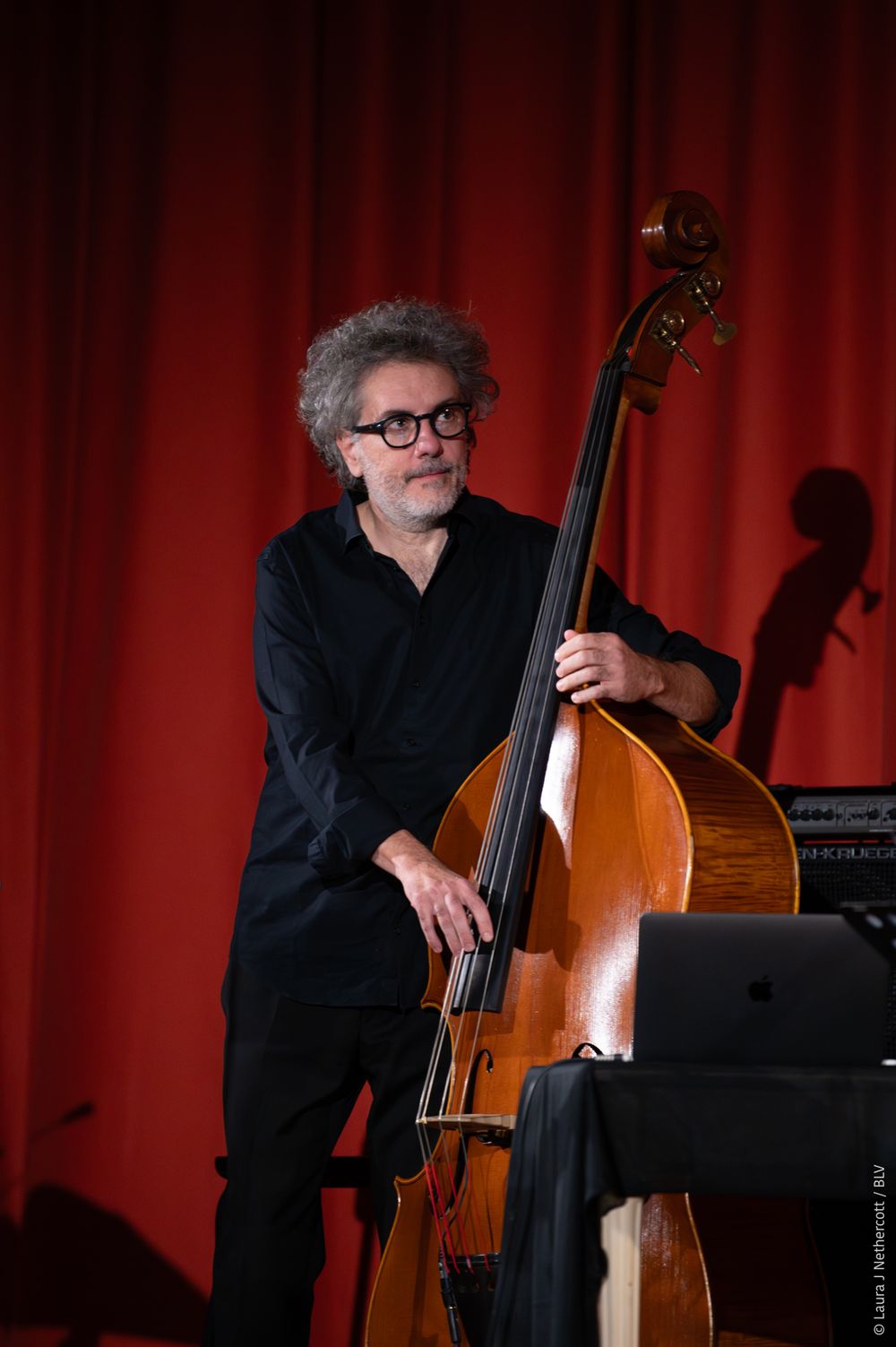 A musician plays upright bass on stage against a red curtain backdrop in dramatic lighting.