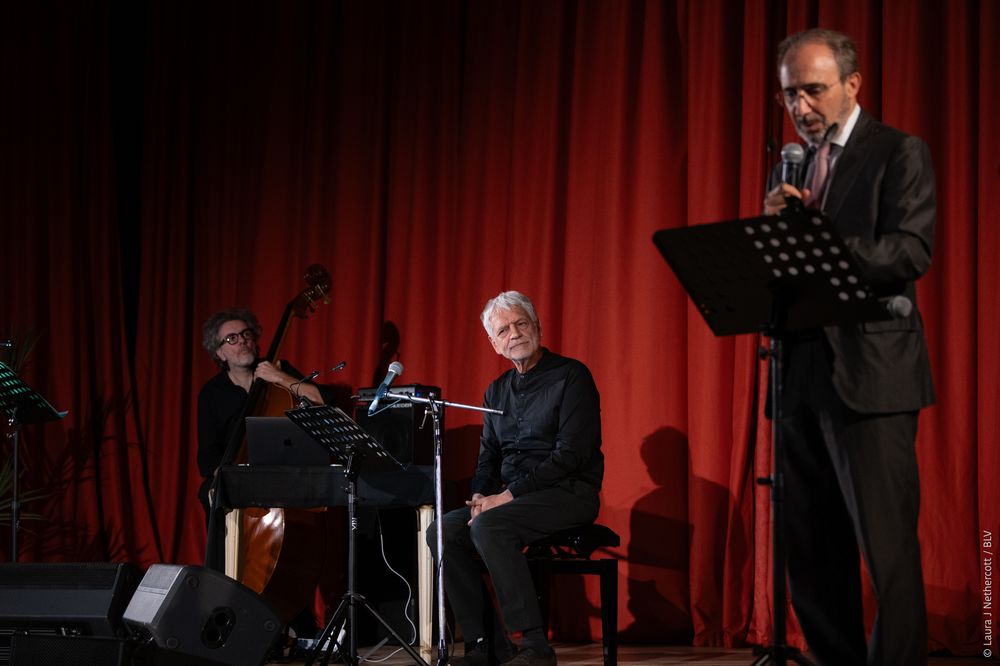 Musicians perform on stage with red curtain backdrop during live performance at concert venue.