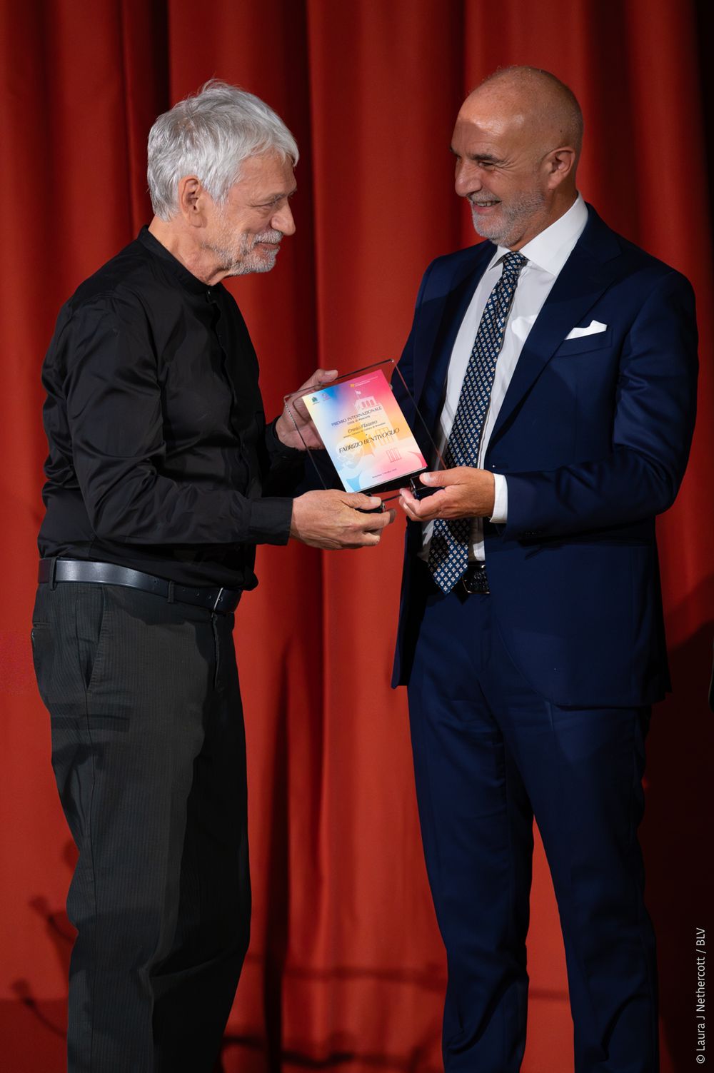 Two men in formal attire exchange small items in front of a red curtain backdrop.