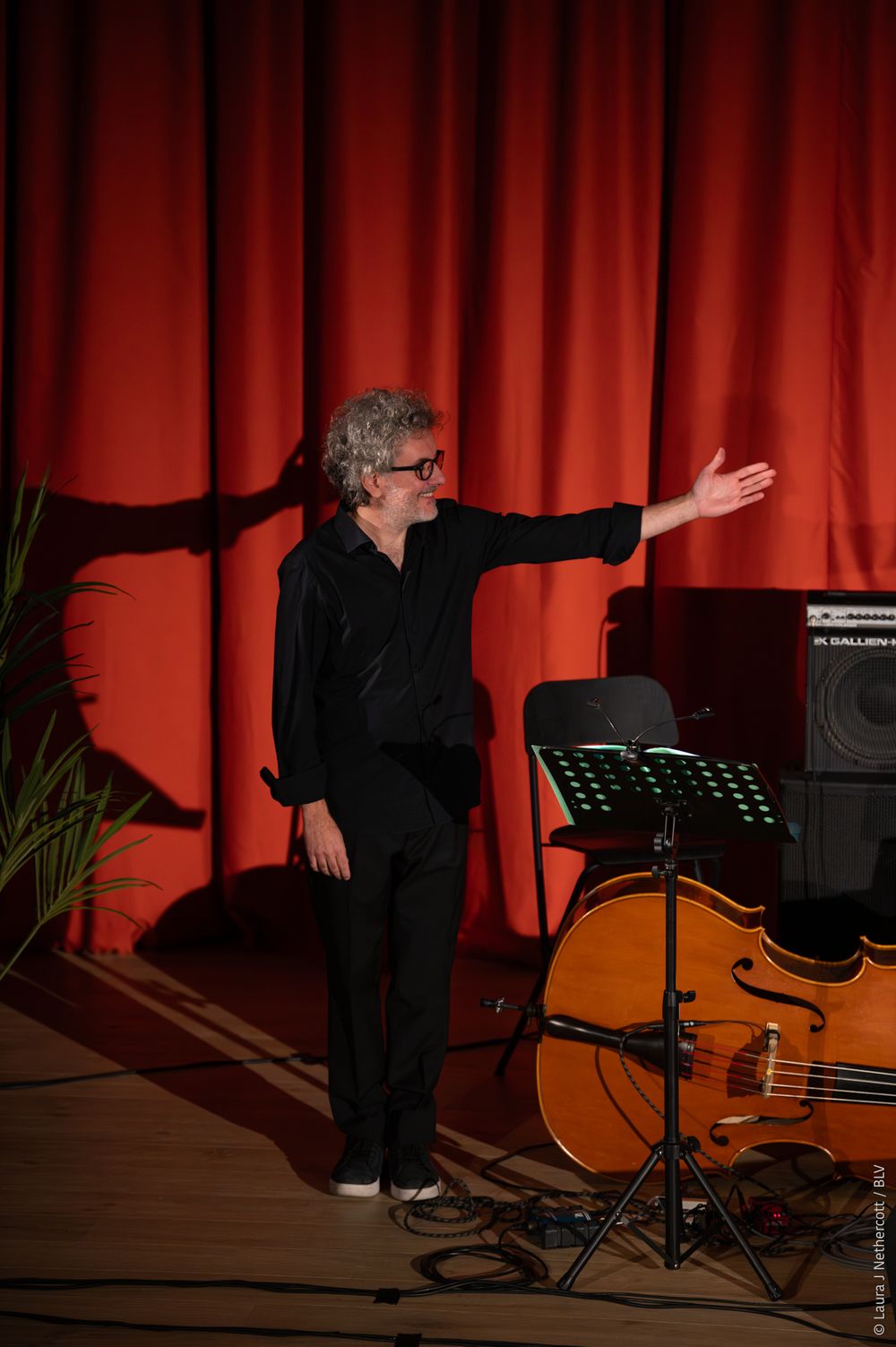A performer in black clothing moves dynamically across a stage with a red curtain backdrop and music stand.