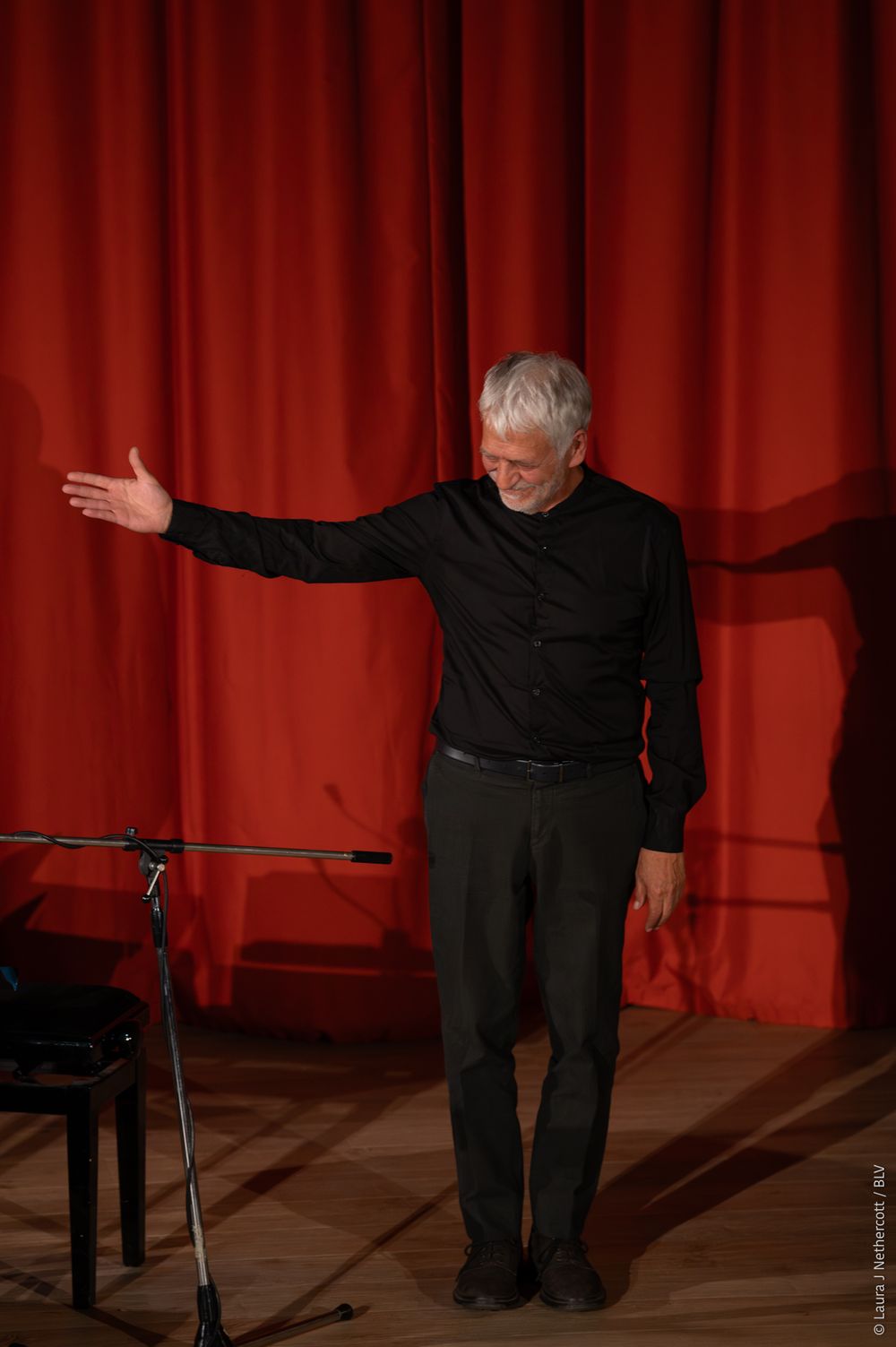 A performer in black attire bows deeply on stage with a red curtain backdrop.