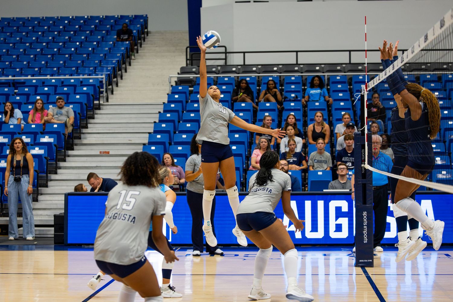 A volleyball player jumps to spike the ball during an indoor match with blue stadium seating in the background.