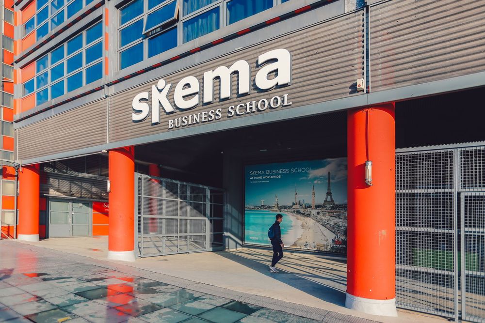 Exterior of SKEMA Business School building with modern architecture, red accents, and a student walking at the entrance.