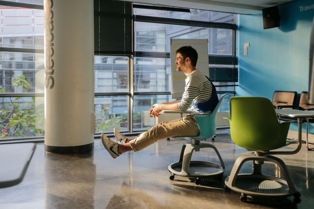 Male student balancing on a modern wobble chair by large windows in a contemporary classroom.
