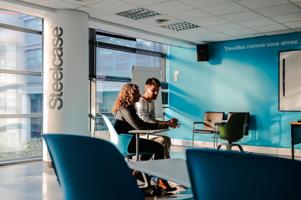 Two students collaborating at a desk in a modern Steelcase-furnished study space with blue walls.