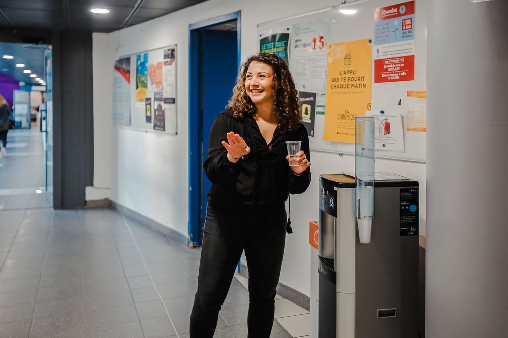 Female student smiling while using a water dispenser in a bright school corridor.