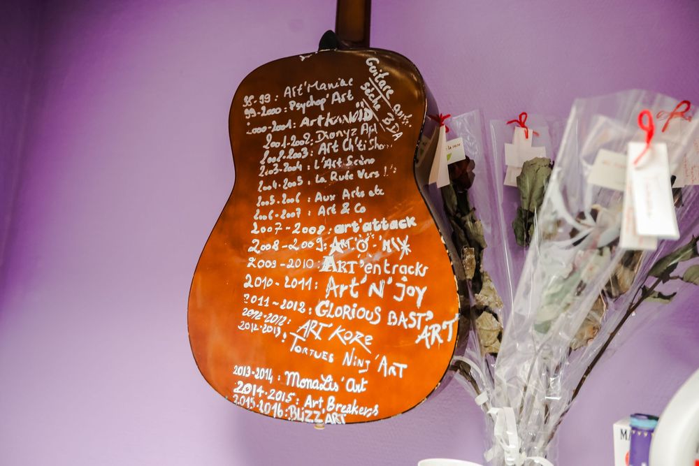 A wooden guitar decorated with handwritten text and dates displayed on a purple wall beside dried flowers.