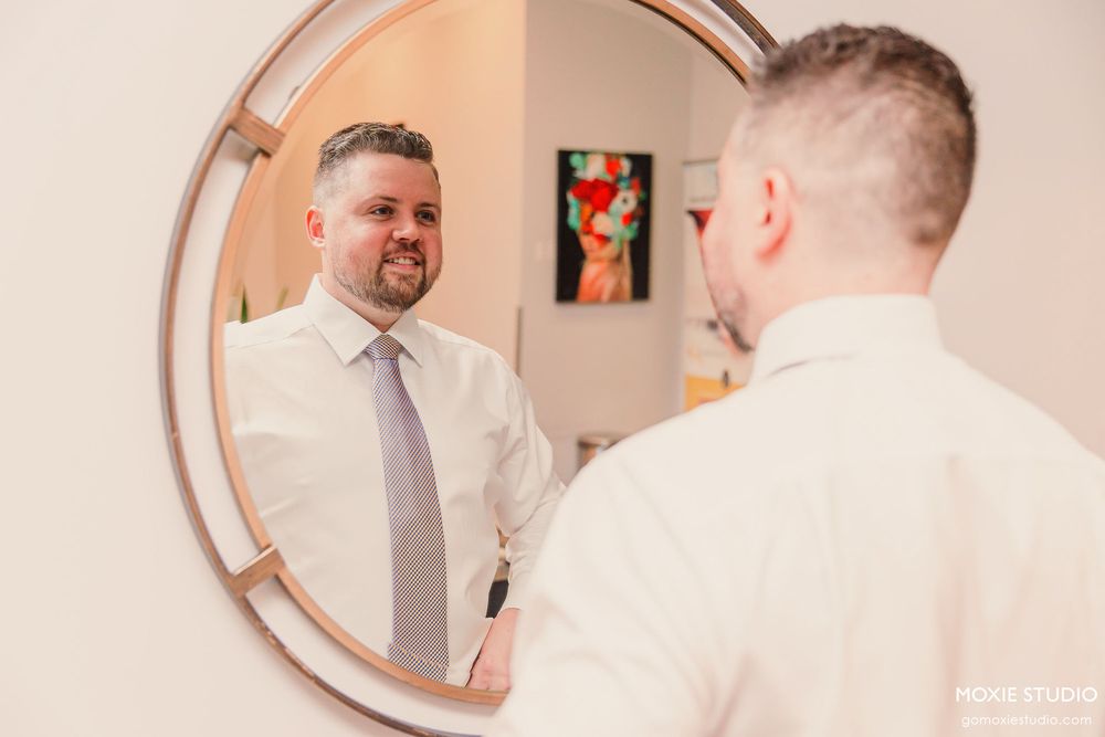 Man in light colored shirt and tie adjusts his appearance in a round copper-framed mirror.