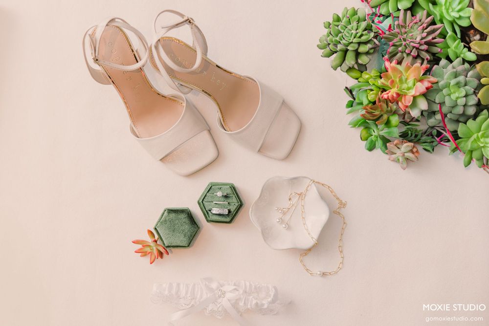 Wedding details including blush pink heels and green succulent decorations arranged on light beige surface.