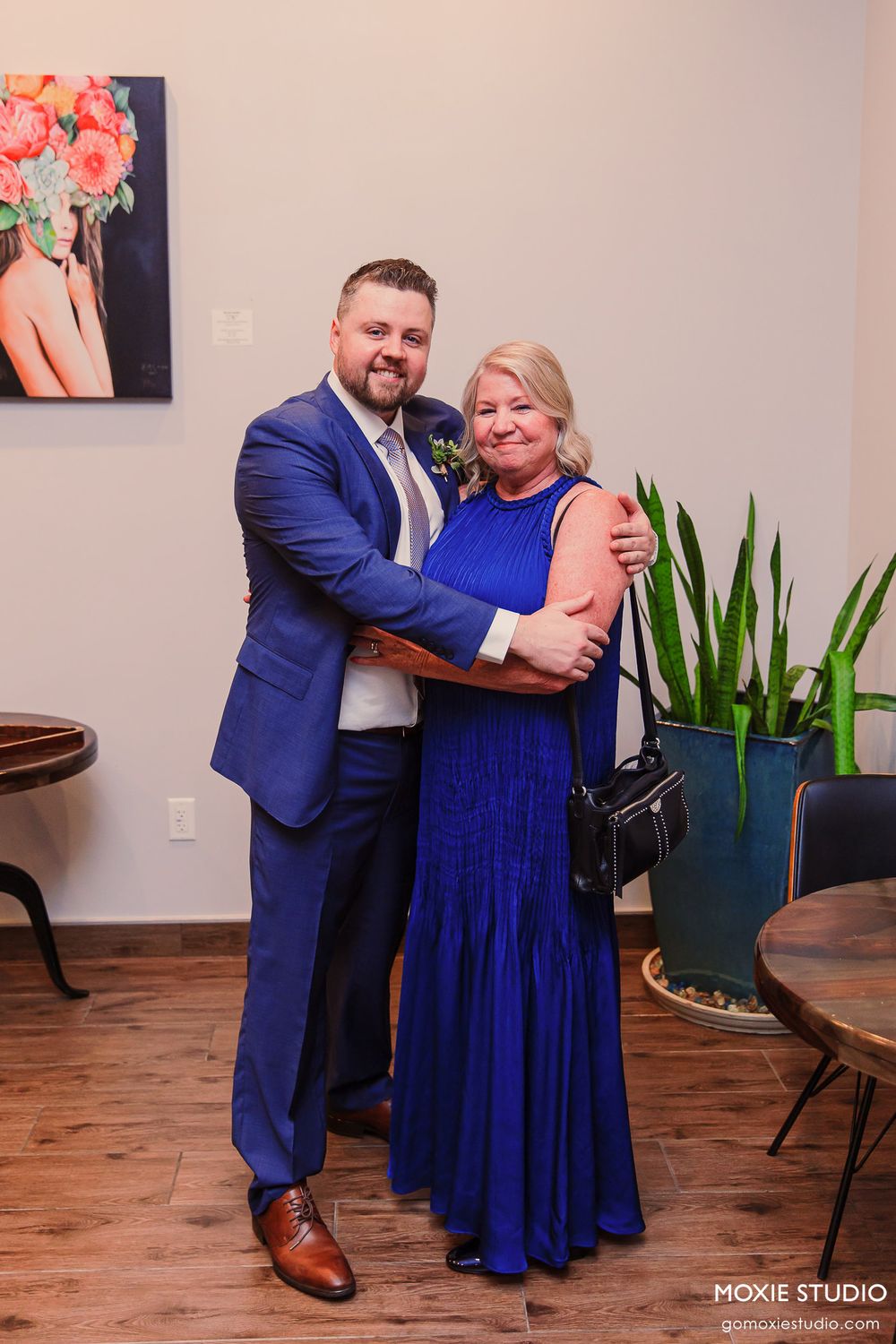 Two people in formal attire, one in a blue suit and one in a royal blue dress, share a joyful moment on a wooden floor.
