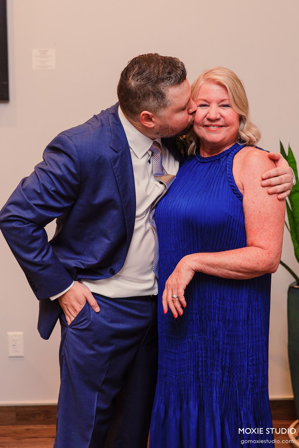 A couple in formal attire shares a tender moment, with the person in a blue dress and another in a navy suit embracing.